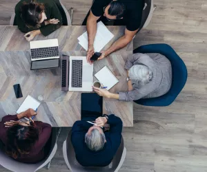 A group of workers are sat at a boardroom table working and conversing.
